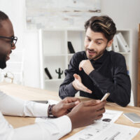 portrait-two-young-smiling-multinational-businessmen-sitting-table-happily-discussing-new-project-while-working-together-office-isolated